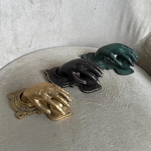 May include: Three decorative hand-shaped door knockers in gold, black, and green. Each knocker features a detailed hand emerging from a decorative base. The knockers are arranged on a light gray surface.
