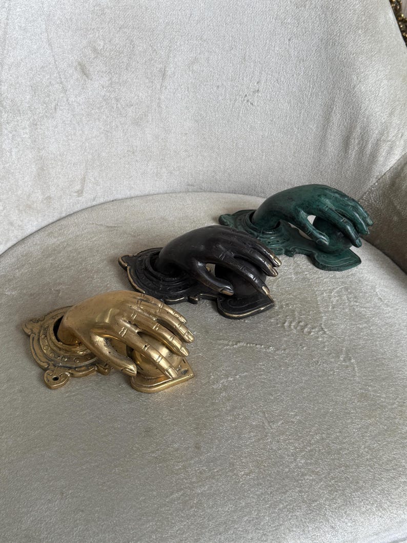 May include: Three decorative hand-shaped door knockers in gold, black, and green. Each knocker features a detailed hand emerging from a decorative base. The knockers are arranged on a light gray surface.