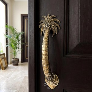 May include: A brass door handle shaped like a palm tree with a textured trunk and detailed fronds, mounted vertically on a dark brown wooden door. The base of the handle features a circular plate with the engraved text SUKO HOME.