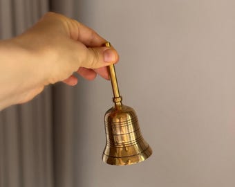 Könnte beinhalten: Eine kleine, goldfarbene Glocke mit langem Griff. Die Glocke hat einen ausgestellten Sockel und horizontale Linien um ihren Körper. Der Griff ist dünn und zylindrisch. Die Glocke wird vor einem neutralen Hintergrund gehalten.