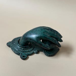 May include: A teal-colored hand-shaped door knocker. The hand emerges from an ornate base with a detailed, aged appearance. The door knocker is made of metal and has a weathered finish, suggesting an antique or vintage style.