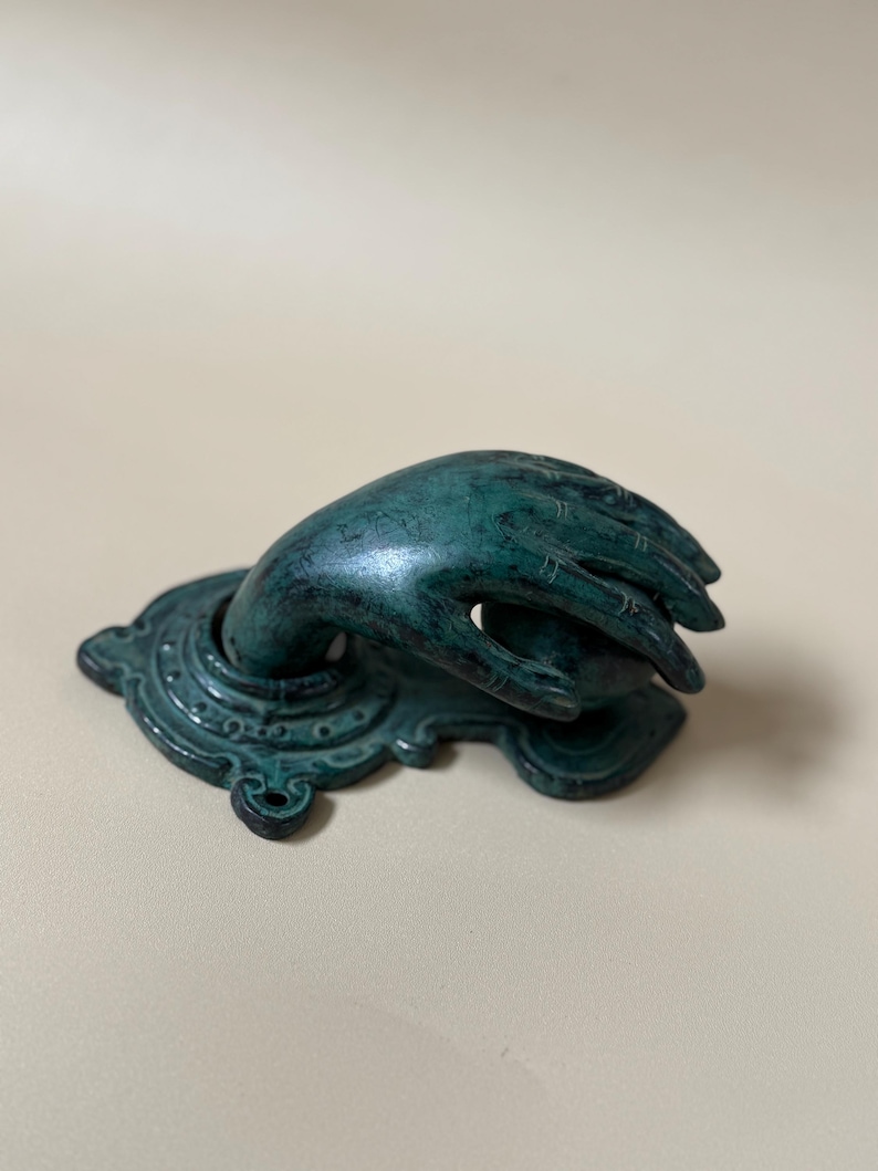 May include: A teal-colored hand-shaped door knocker. The hand emerges from an ornate base with a detailed, aged appearance. The door knocker is made of metal and has a weathered finish, suggesting an antique or vintage style.