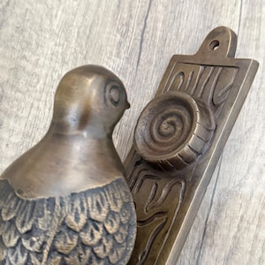 May include: A bronze-toned metal door knocker featuring a bird figure attached to a rectangular backplate. The backplate displays a raised circular spiral design and a top mounting hole. The bird has detailed feather textures on its body.