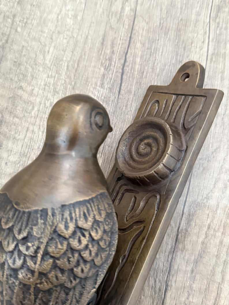 May include: A bronze-toned metal door knocker featuring a bird figure attached to a rectangular backplate. The backplate displays a raised circular spiral design and a top mounting hole. The bird has detailed feather textures on its body.