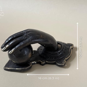 May include: A bronze-toned door knocker shaped like a hand reaching for a sphere. The decorative piece is 6.3 inches wide and 3.9 inches tall. The hand is detailed with visible knuckles and fingernails. The base has an ornate design.