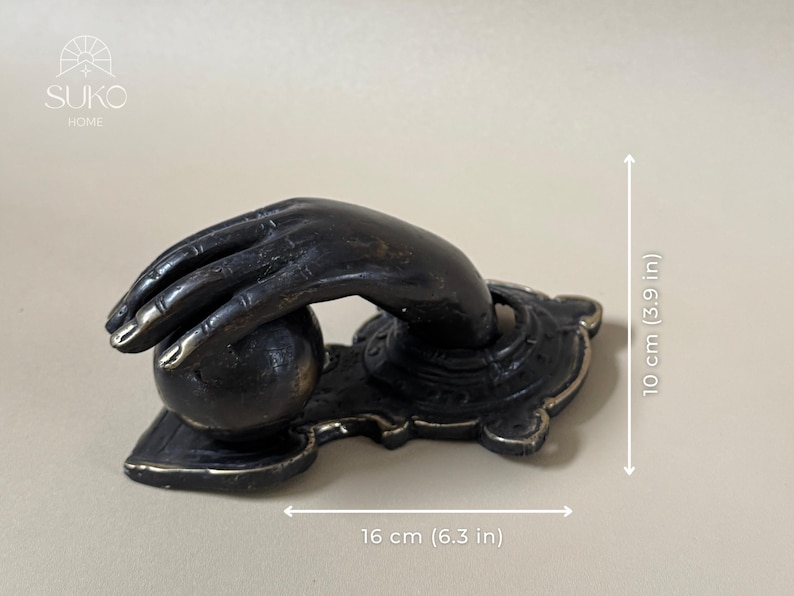 May include: A bronze-toned door knocker shaped like a hand reaching for a sphere. The decorative piece is 6.3 inches wide and 3.9 inches tall. The hand is detailed with visible knuckles and fingernails. The base has an ornate design.