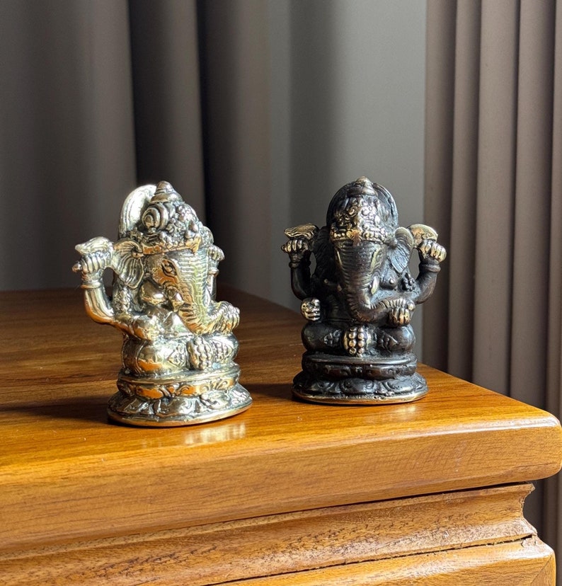 May include: Two small, ornate, brass elephant statues. One is gold and the other is dark brown. They are both sitting on a wooden surface.
