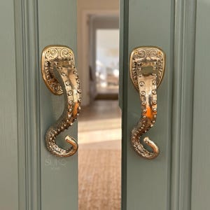 May include: Two metallic gold-toned door handles shaped like octopus tentacles, featuring detailed suction cups and ornate mounting plates. The handles are attached to sage green double doors, with the text SUKO HOME visible on the left.