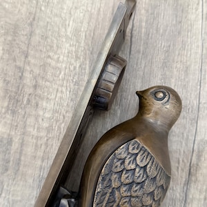 May include: A bronze-toned metal door knocker featuring a bird figure with detailed wing feathers attached to a vertical mounting plate. The bird is positioned at the bottom of the plate, which includes a circular hinge mechanism at the top.