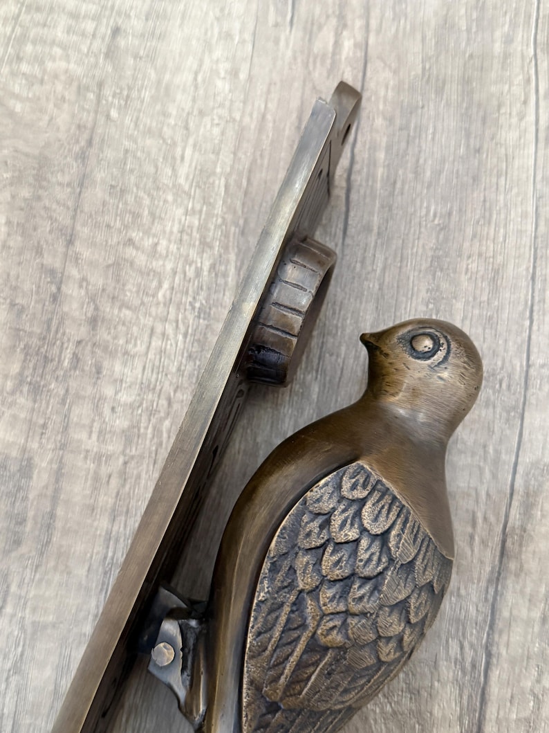 May include: A bronze-toned metal door knocker featuring a bird figure with detailed wing feathers attached to a vertical mounting plate. The bird is positioned at the bottom of the plate, which includes a circular hinge mechanism at the top.