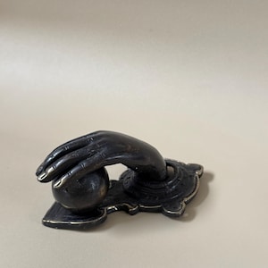 May include: Bronze-toned door knocker featuring a hand reaching for a sphere. The hand is detailed with visible knuckles and fingernails. The knocker is mounted on a decorative base with an ornate design, suitable for a front door.