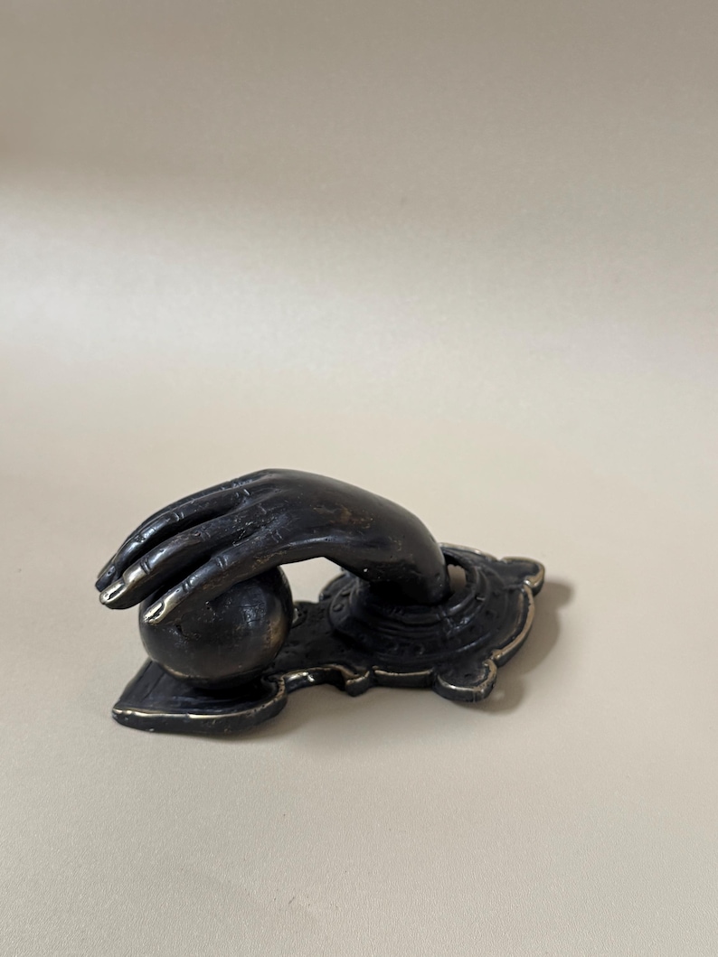 May include: Bronze-toned door knocker featuring a hand reaching for a sphere. The hand is detailed with visible knuckles and fingernails. The knocker is mounted on a decorative base with an ornate design, suitable for a front door.