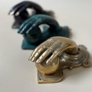 May include: Three decorative door knockers in a row. The front knocker is gold-toned, with a hand grasping a sphere. The other two knockers are in the background, one is teal and the other is black.