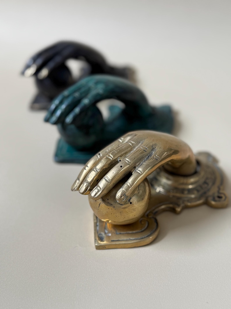 May include: Three decorative door knockers in a row. The front knocker is gold-toned, with a hand grasping a sphere. The other two knockers are in the background, one is teal and the other is black.