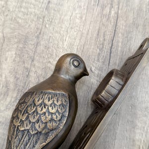 May include: A bronze-toned metal door knocker featuring a sculpted bird with detailed wing feathers. The bird is attached to a rectangular backplate that includes a circular striking plate and a mounting hole at the top for installation on a door.