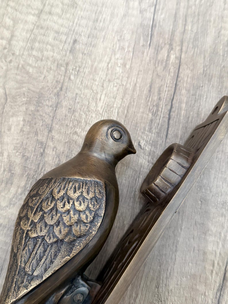 May include: A bronze-toned metal door knocker featuring a sculpted bird with detailed wing feathers. The bird is attached to a rectangular backplate that includes a circular striking plate and a mounting hole at the top for installation on a door.