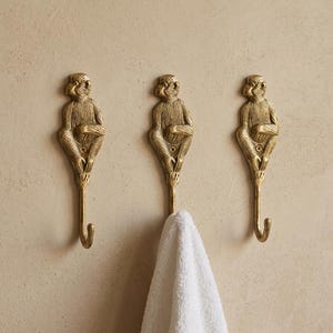 May include: Three brass monkey-shaped wall hooks. Each hook features a seated monkey figure with a curved hook extending downwards. A white towel hangs from one hook, set against a textured beige wall.