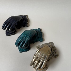 May include: Three decorative hand-shaped door knockers in black, teal, and gold. Each knocker features a detailed hand design with a decorative backplate. The gold knocker has a heart-shaped base.