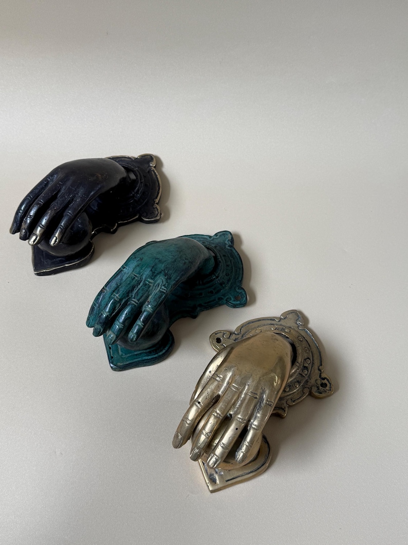 May include: Three decorative hand-shaped door knockers in black, teal, and gold. Each knocker features a detailed hand design with a decorative backplate. The gold knocker has a heart-shaped base.