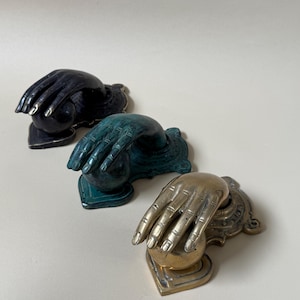 May include: Three decorative door knockers in a row. The knockers are shaped like hands. The first is black with gold accents, the second is teal, and the third is brass-colored.