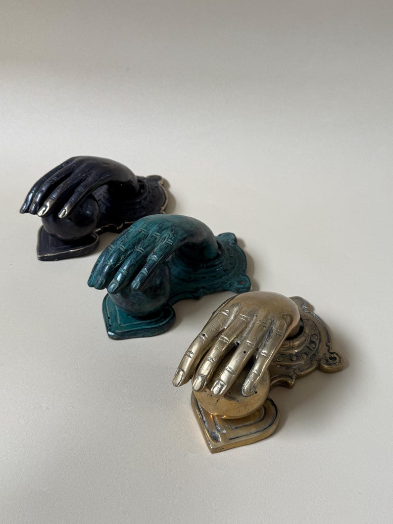 May include: Three decorative door knockers in a row. The knockers are shaped like hands. The first is black with gold accents, the second is teal, and the third is brass-colored.