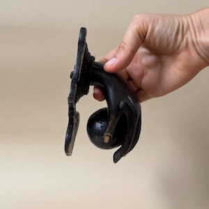 May include: A dark bronze door knocker in the shape of a hand holding a round ball. The hand is detailed with visible knuckles and a slightly curved shape. The knocker is mounted on a decorative rectangular plate.