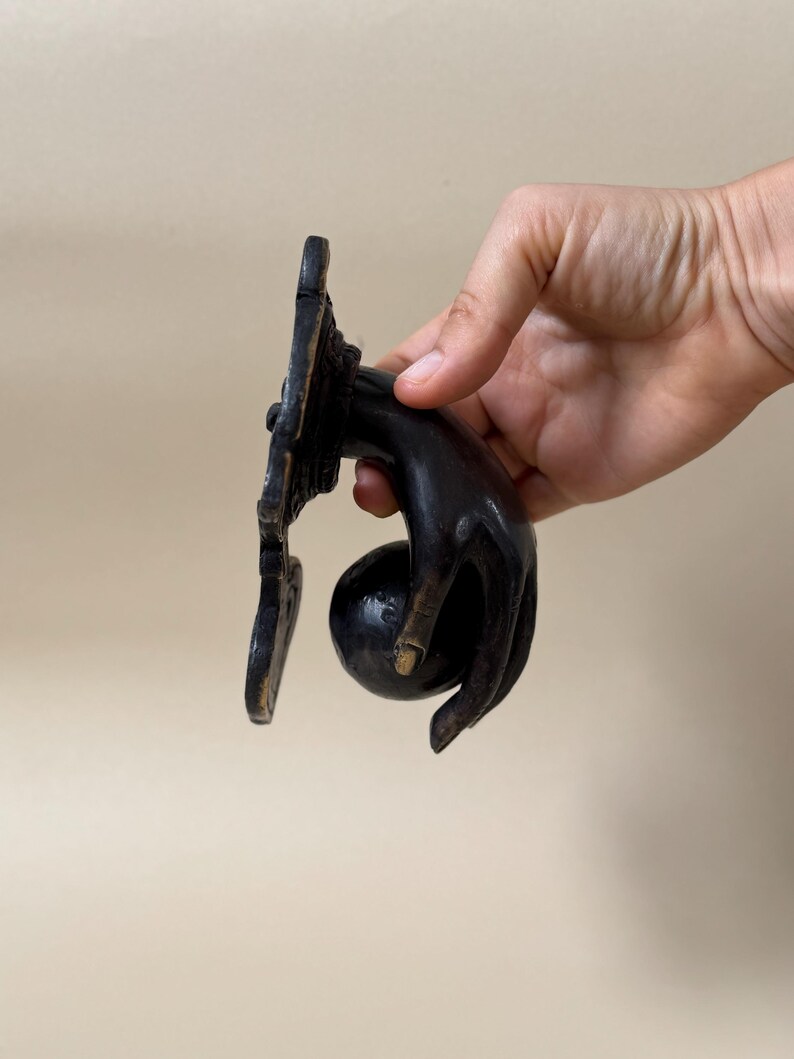 May include: A dark bronze door knocker in the shape of a hand holding a round ball. The hand is detailed with visible knuckles and a slightly curved shape. The knocker is mounted on a decorative rectangular plate.
