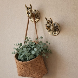 Puede incluir: Dos ganchos de pared de metal dorado con forma de manos haciendo un gesto de rock-and-roll, montados sobre bases circulares. Una cesta de cuero beige trenzado con una correa larga cuelga de un gancho, conteniendo una pequeña planta verde.