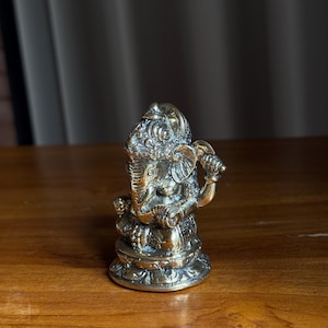 May include: A gold-colored brass statue of Ganesha, the Hindu god of wisdom and prosperity, sitting on a lotus base. The statue is intricately detailed and has a traditional design.