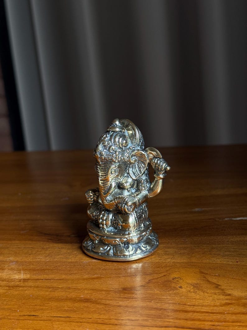 May include: A gold-colored brass statue of Ganesha, the Hindu god of wisdom and prosperity, sitting on a lotus base. The statue is intricately detailed and has a traditional design.