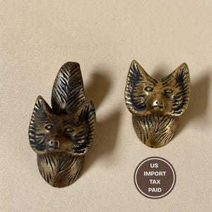 Może przedstawiać: Dwa mosiężne uchwyty do szafek w kształcie głowy lisa. Jeden uchwyt ma wzór przypominający pióro. Antyczne wykończenie. Tekst "US IMPORT TAX PAID" znajduje się w okręgu. Ozdobne okucia meblowe.