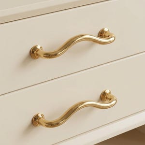 Può includere: Primo piano di due cassetti color crema con maniglie dorate e curve. Le maniglie hanno un design a onda e sono attaccate ai cassetti con supporti circolari. I cassetti fanno parte di un mobile più grande.