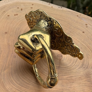 May include: A brass door knocker in the shape of a fist, with an ornate backplate. The knocker has a polished finish and a curved handle. The design is detailed and vintage-inspired, suitable for a front door.
