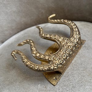 May include: A brass-coloured wall hook in the shape of an octopus tentacle. The hook has a textured surface with small, circular details and a diamond-shaped base. The tentacles curve upwards.