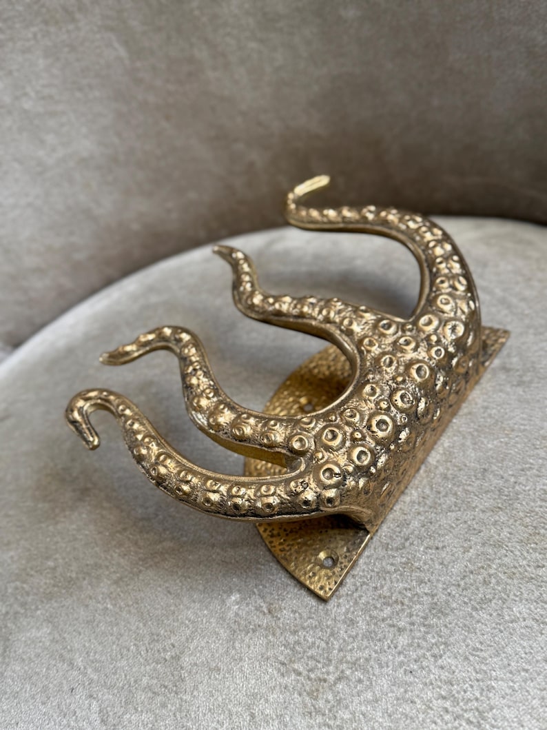 May include: A brass-coloured wall hook in the shape of an octopus tentacle. The hook has a textured surface with small, circular details and a diamond-shaped base. The tentacles curve upwards.