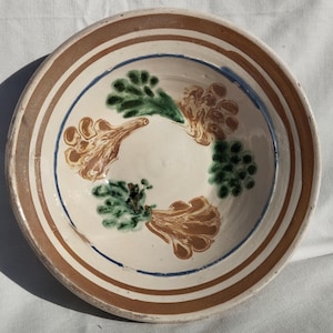 May include: A ceramic bowl with a cream-colored interior and a brown and blue rim. The bowl is decorated with green and brown floral designs. The bowl is likely used for serving food.