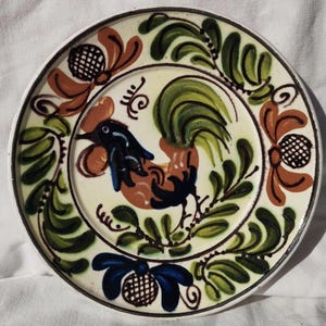 May include: A hand-painted ceramic plate with a rooster design. The central image is a rooster in orange, blue, and black, with a green tail. The plate is decorated with floral and leaf patterns in green and brown, with a white background and a black rim.