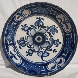 May include: Antique ceramic plate with a white center and a blue floral design. The plate's border is blue with a decorative pattern. The plate has a dark rim and is likely vintage.