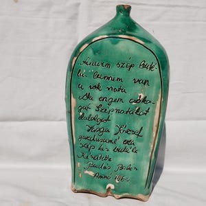 Może przedstawiać: Zielona ceramiczna butelka z wąską szyjką i zaokrąglonym korpusem. Butelka ma ciemnozieloną obwódkę i odręczny tekst czarnym tuszem. Tekst wydaje się być wierszem lub inskrypcją. Butelka jest przedmiotem dekoracyjnym.
