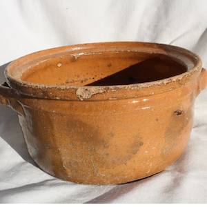 Puede incluir: Una maceta grande, redonda, de terracota con dos asas. La maceta está vidriada y tiene un color marrón claro. Tiene un aspecto desgastado y rústico.