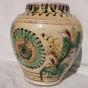 Op de afbeelding: Een handbeschilderde keramische vaas met een crèmekleurige basis. De vaas heeft groene, gele en bruine bloemmotieven. De rand heeft een decoratief patroon. De vaas is ongeveer 25 cm hoog.