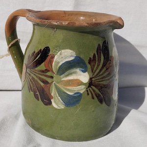 Może przedstawiać: Zielony ceramiczny dzbanek z ręcznie malowanym motywem kwiatowym w odcieniach bieli, błękitu i ciemnego brązu. Naczynie ma szeroki, zaokrąglony korpus, zakrzywiony uchwyt oraz mały dzióbek przy krawędzi. Gliniana faktura jest widoczna na uchwycie.