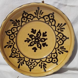 May include: A round, yellow ceramic plate with a scalloped edge. The plate features a black, symmetrical floral design in the center and around the rim. The plate is likely handmade and has a rustic aesthetic.