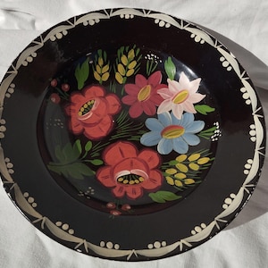 Può includere: Un piatto decorativo rotondo e nero con un motivo floreale dipinto a mano. I fiori sono rossi, rosa e blu, con foglie verdi e dettagli gialli. Il bordo ha un bordo bianco smerlato.
