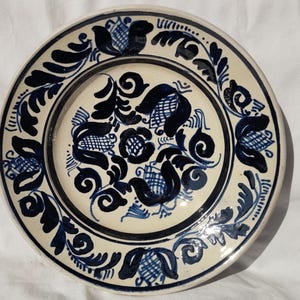 May include: A round, off-white ceramic plate with a hand-painted floral design in shades of blue. The plate features a central floral motif surrounded by a border of stylized leaves and flowers. The rim is also decorated with blue patterns.