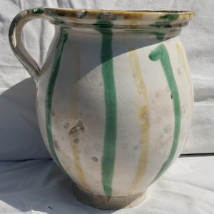 May include: A ceramic pitcher with a white glazed finish featuring vertical stripes in alternating shades of green and yellow. The vessel has a rounded body, a wide mouth, and a single curved handle attached to the side of the upper rim.