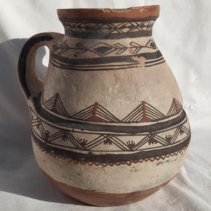 Puede incluir: Jarra de cerámica beige hecha a mano con asa, con patrones geométricos negros y marrones alrededor del cuerpo y el borde. La jarra tiene forma redondeada y base ancha, con borde y base marrón oscuro.