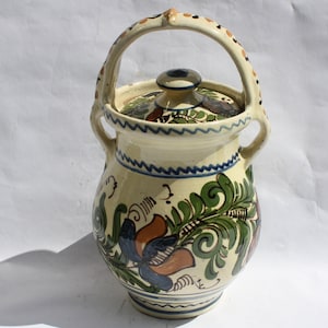 May include: A white ceramic jar with a lid and a handle. The jar is decorated with blue, green, and orange floral designs.