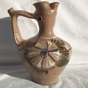 Könnte beinhalten: Handgefertigter, brauner Keramikkrug mit Henkel und Ausguss. Der Krug ist mit einem großen Blumendesign in Blau, Weiß und Grün verziert. Der Krug hat einen bauchigen Körper und einen schmalen Hals.