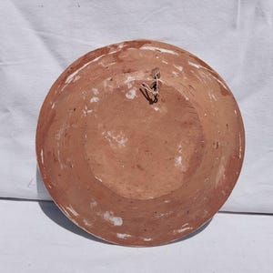 Op de afbeelding: Een ronde terracotta schaal met een roodbruine afwerking en zichtbare witte verfresten op het oppervlak. De achterkant van de schaal is voorzien van een klein metalen draadlusje nabij het midden, bedoeld om het object aan op te hangen.